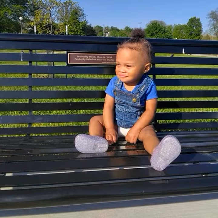 Terri'Jo is registered to the contest to win money with this photo: toddler, child, bench, outdoor, park, grass, trees, blue_sky, sunny, smiling, sitting, clothing, socks, daylight, nature, happy, casual, portrait, social_media, music_overlay