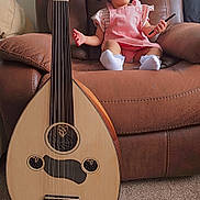 Ayla is registered to the contest to win money with this photo: toddler, child, pink_overalls, white_socks, brown_armchair, musical_instrument, oud, carpet, indoor, window, curious_expression, sitting, holding_object, string_instrument, furniture, home, person, hair_tied, light, cozy