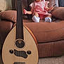 toddler, child, pink_overalls, white_socks, brown_armchair, musical_instrument, oud, carpet, indoor, window, curious_expression, sitting, holding_object, string_instrument, furniture, home, person, hair_tied, light, cozy