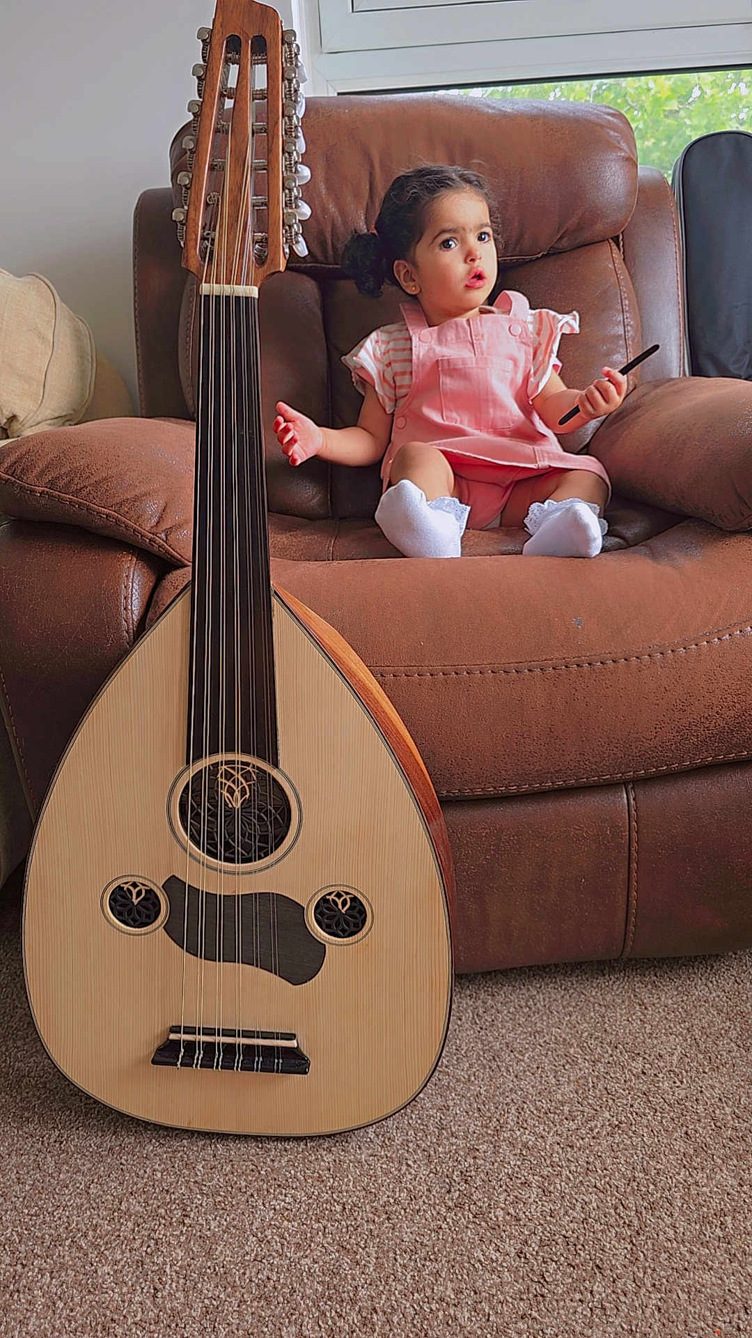 Ayla is registered to the contest to win money with this photo: toddler, child, pink_overalls, white_socks, brown_armchair, musical_instrument, oud, carpet, indoor, window, curious_expression, sitting, holding_object, string_instrument, furniture, home, person, hair_tied, light, cozy