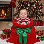 baby, candy_canes, celebration, child, christmas_presents, christmas_tree, decorations, festive_clothing, fireplace, gift_box, green_bow, holiday_decor, holiday_photo, holiday_season, indoors, joyful, red_velvet_sack, smiling, stocking, warm_lighting