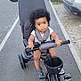 baby, car, child, curious, curly_hair, face, greenery, handlebars, outdoor, pavement, person, road, sandals, seatbelt, sidewalk, stroller, toddler, transportation, tricycle, vehicle
