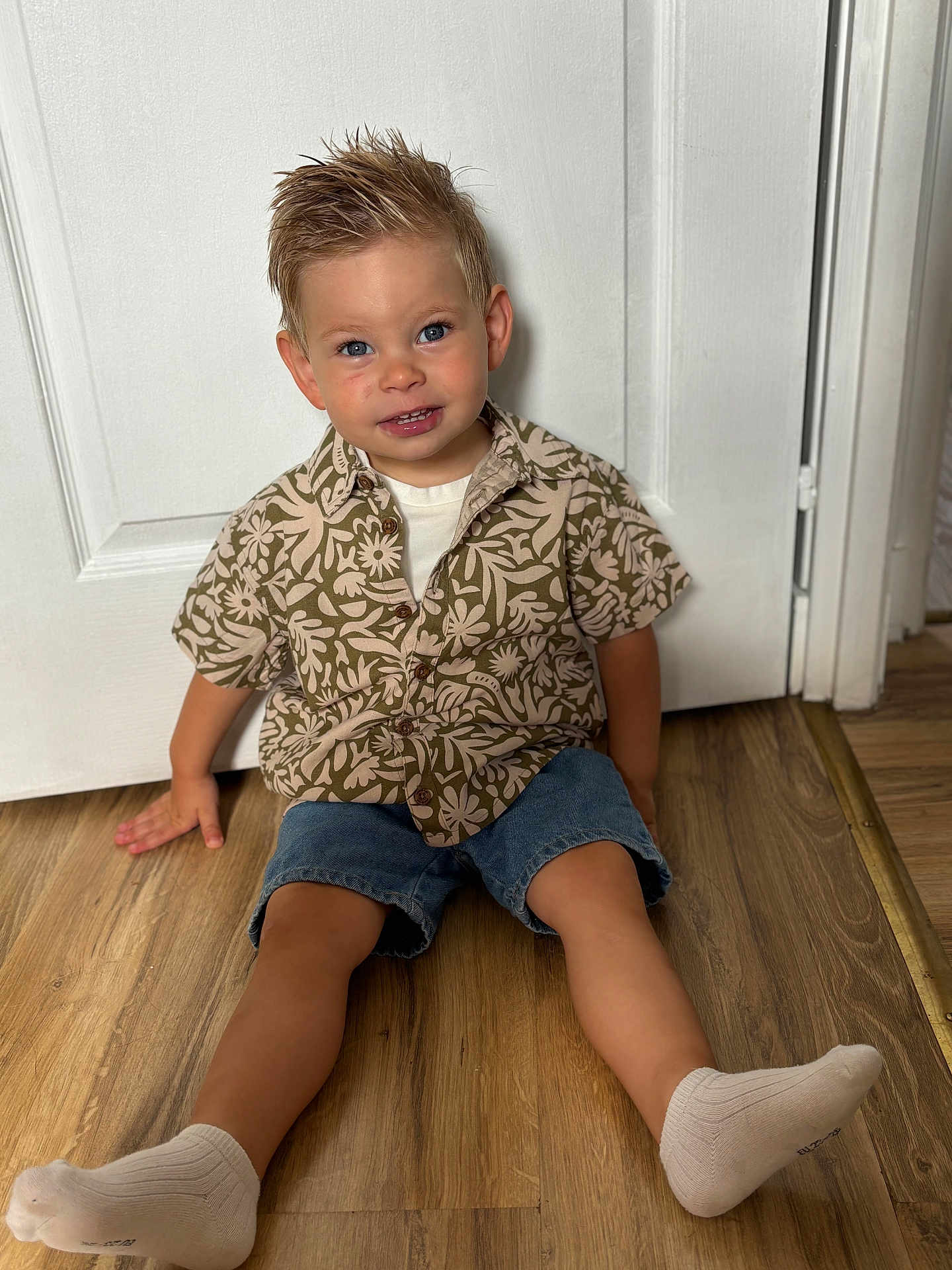 Charly participe au concours pour gagner de l'argent avec cette photo : blue_eyes, boy, casual_clothing, child, cute, denim_shorts, door, floor, hair, happy, home, indoor, portrait, shirt, sitting, smiling, socks, toddler, wooden_floor, young_child