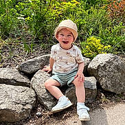 Charly a rejoint le concours — aidez-le/la à gagner de superbes lots ! toddler, child, hat, smiling, rocks, greenery, outdoor, sunlight, shorts, white_shoes, nature, plants, happy, sitting, casual_clothing, daylight, portrait, young_child, trees, ground