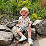 toddler, child, hat, smiling, rocks, greenery, outdoor, sunlight, shorts, white_shoes, nature, plants, happy, sitting, casual_clothing, daylight, portrait, young_child, trees, ground