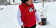 Arturo is registered to the contest to win money with this photo: child, toddler, snow, winter, beanie, hat, red_vest, mickey_mouse, outdoor, trees, house, potted_plant, boots, footprints, standing, jacket, snowfall, yard, curious, portrait