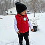 child, toddler, snow, winter, beanie, hat, red_vest, mickey_mouse, outdoor, trees, house, potted_plant, boots, footprints, standing, jacket, snowfall, yard, curious, portrait