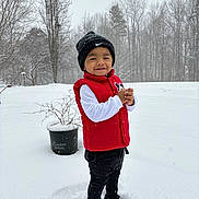 Arturo is registered to the contest to win money with this photo: child, toddler, snow, winter, beanie, hat, red_vest, white_long_sleeve, black_pants, boots, smile, outdoors, trees, potted_plant, snowflakes, standing, portrait, playful, backyard, cold_weather