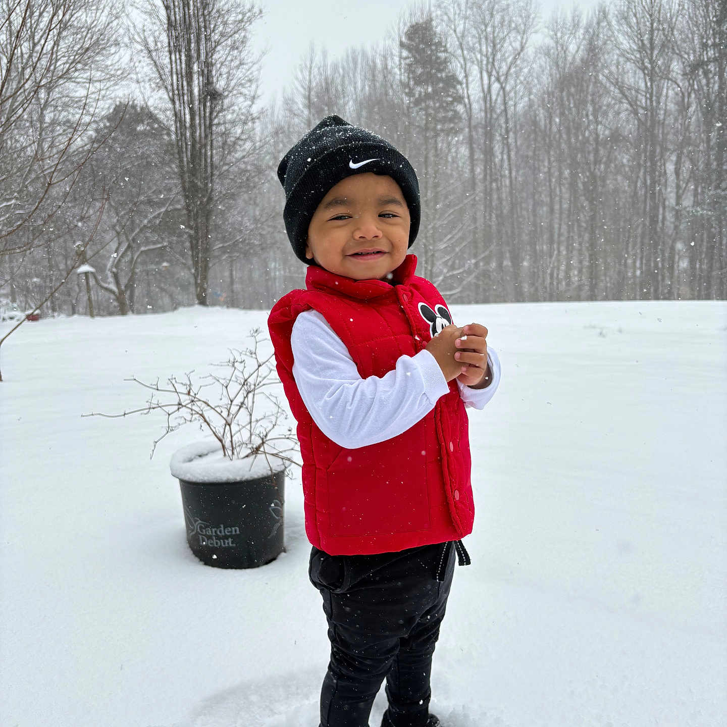 Arturo is registered to the contest to win money with this photo: backyard, beanie, black_pants, boots, child, cold_weather, hat, outdoors, playful, portrait, potted_plant, red_vest, smile, snow, snowflakes, standing, toddler, trees, white_long_sleeve, winter
