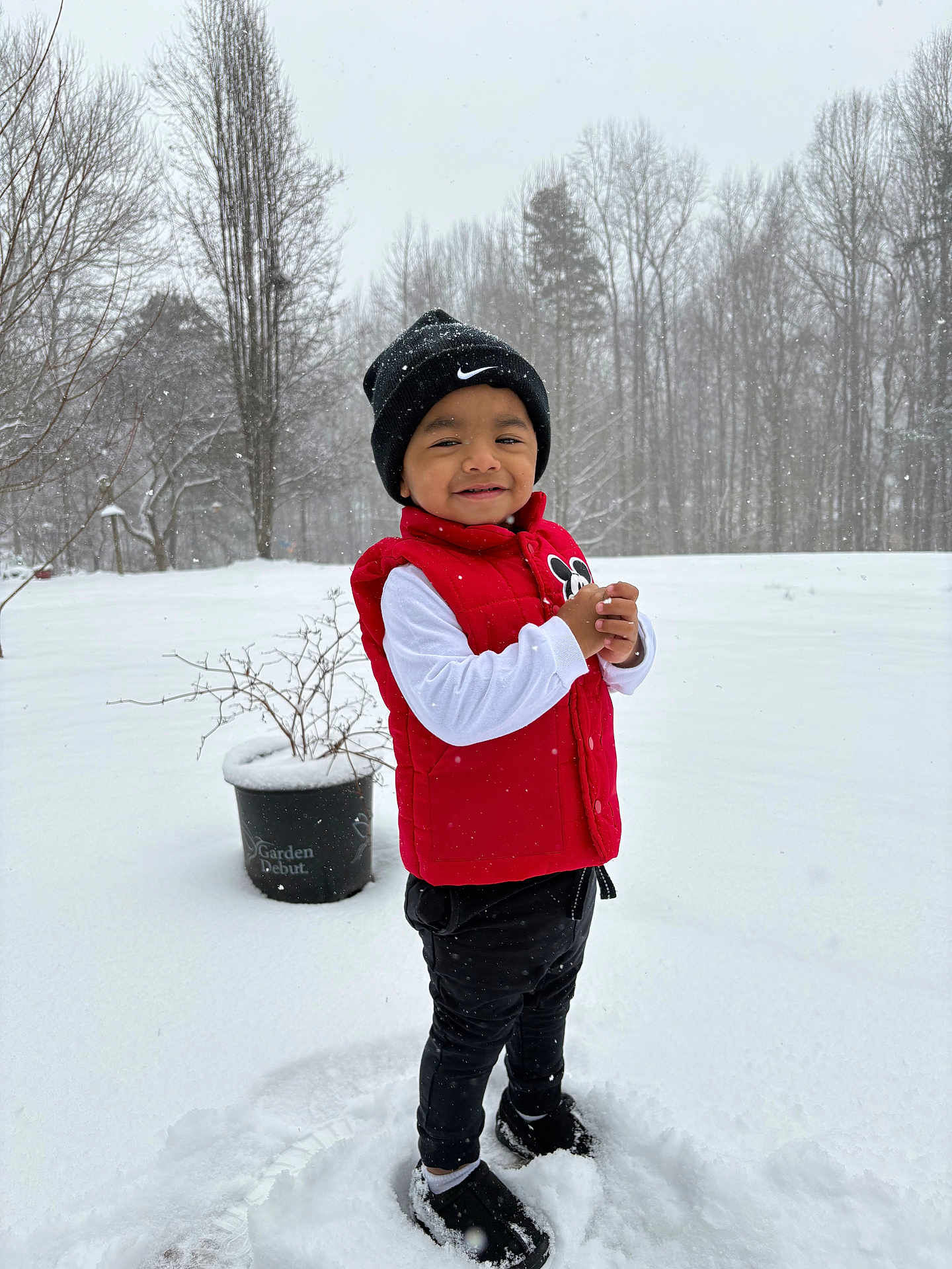Arturo is registered to the contest to win money with this photo: child, toddler, snow, winter, beanie, hat, red_vest, white_long_sleeve, black_pants, boots, smile, outdoors, trees, potted_plant, snowflakes, standing, portrait, playful, backyard, cold_weather