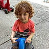 child, curly_hair, outdoor, playground, ride_on_toy, toy_vehicle, blue_toy, red_toy, sitting, portrait, looking_at_camera, casual_clothing, orange_shirt, gray_shorts, sneakers, pavement, daylight, playtime, happy, young_person