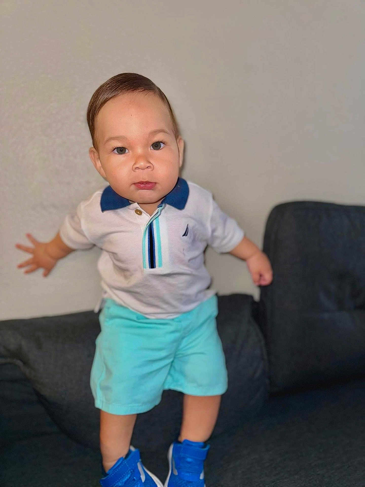 Ethan is registered to the contest to win money with this photo: toddler, child, boy, standing, white_polo, blue_shoes, turquoise_shorts, couch, indoor, furniture, person, young_child, confident, portrait, casual_clothing, cute, home, smiling, face, skin