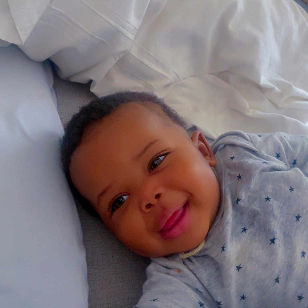 Mouhamed a rejoint le concours — aidez-le/la à gagner de superbes lots ! arm, baby, bed, blanket, child, cute, face, happy, indoor, infant, lying_down, onesie, person, pillow, resting, skin, sleepwear, smiling, star_pattern, young
