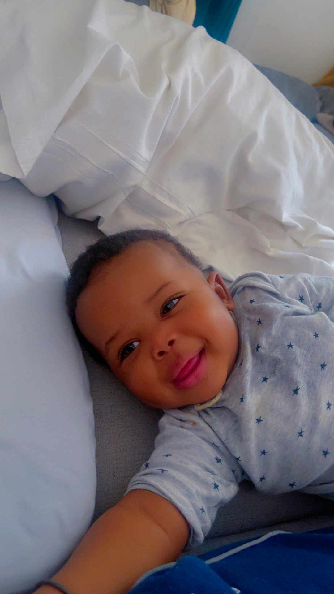 Mouhamed a rejoint le concours — aidez-le/la à gagner de superbes lots ! baby, child, smiling, bed, pillow, blanket, onesie, star_pattern, infant, cute, happy, face, skin, arm, sleepwear, indoor, resting, lying_down, young, person