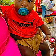 Legend joined the competition — help win amazing prizes! baby, child, pacifier, red_shirt, puma_logo, shorts, sitting, person, outdoor, greenery, people, social_media, mobile_screen, lap, curious, casual, daylight, face, black_hair, expression