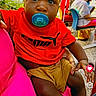 baby, child, pacifier, red_shirt, puma_logo, shorts, sitting, person, outdoor, greenery, people, social_media, mobile_screen, lap, curious, casual, daylight, face, black_hair, expression