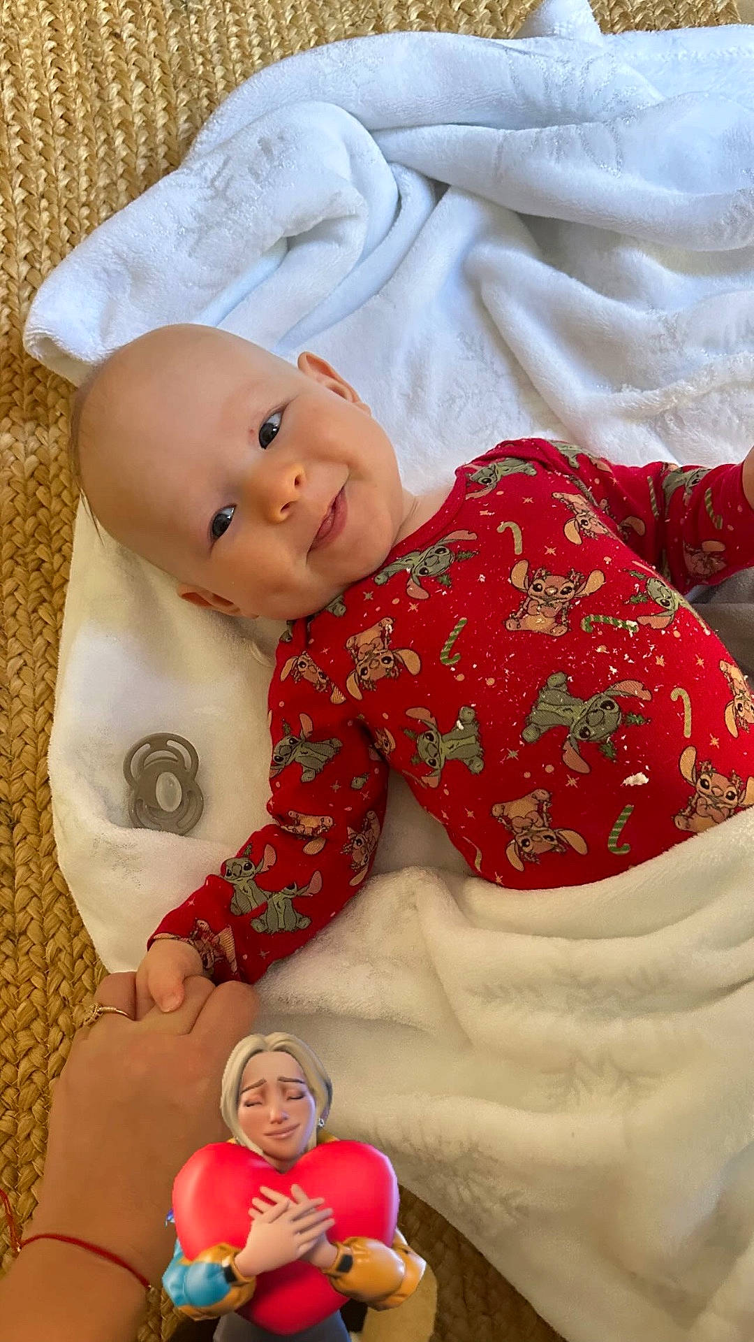 adult_hand, animation, baby, cartoon_print, child, comfort, cozy, cute, hand_holding, happy, heart, indoor, infant, love, person, portrait, red_onesie, smiling, white_blanket, woven_mat