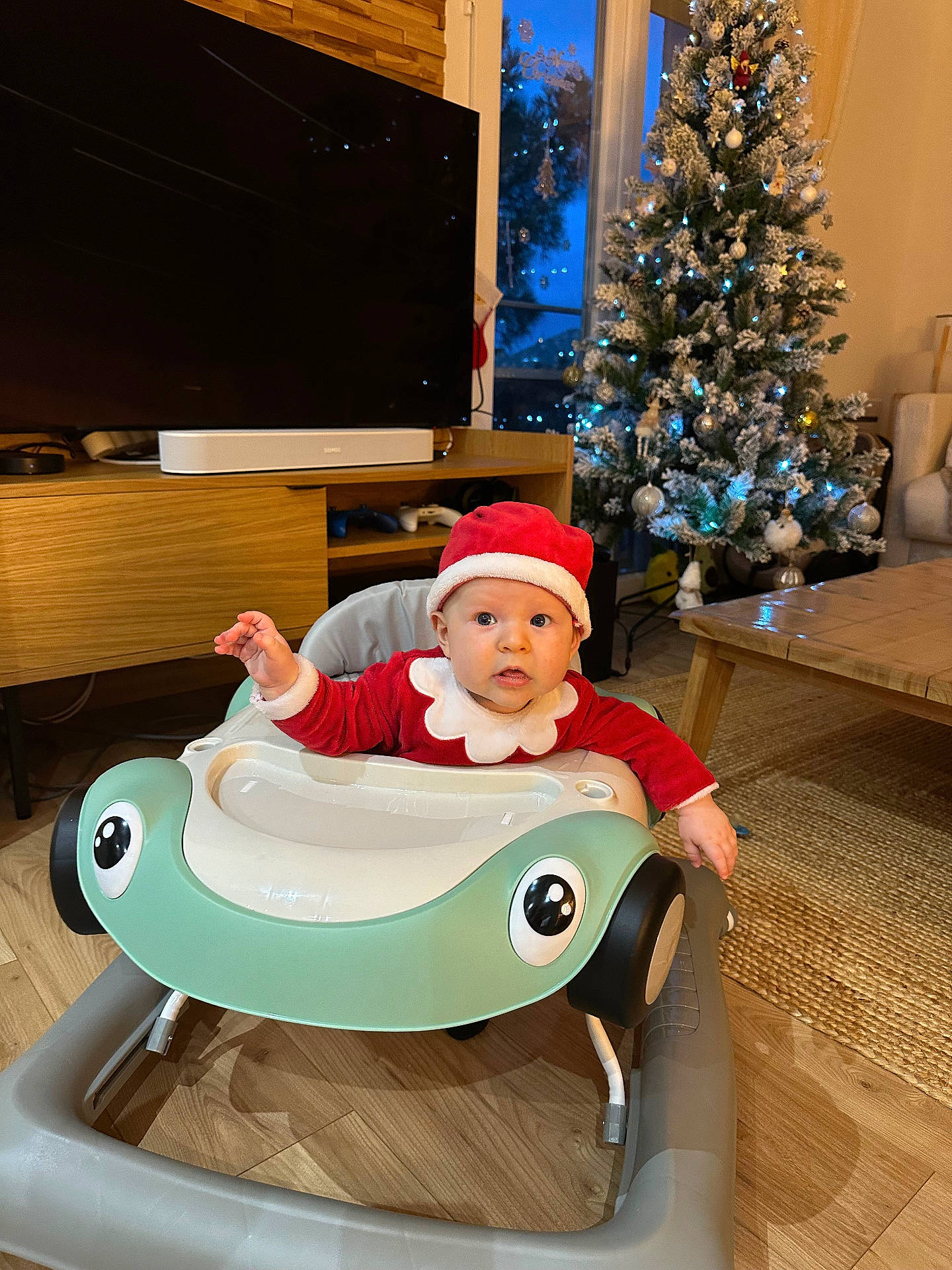 baby, child, christmas_lights, christmas_tree, cute, decorations, festive, furniture, holiday, holiday_season, indoors, living_room, person, santa_hat, smiling, table, television, toy, walker, wood_floor