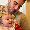 baby, man, child, smiling, crown, red_sweater, bib, happy, family, indoor, table, hands, beard, portrait, clothing, person, affection, celebration, headwear, tablecloth