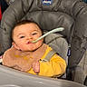 baby, child, high_chair, spoon, bib, messy, eating, indoor, face, expression, yellow_clothing, toy, feeding_time, cheeky, seatbelt, plastic, plaything, home, infant, smile