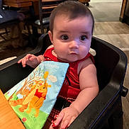 Ophelia joined the competition — help win amazing prizes! baby, child, high_chair, red_clothing, book, winnie_the_pooh, indoor, restaurant, table, wooden_table, person, cute, seated, toy, colorful, infant, face, arm, hand, furniture