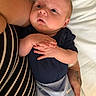 baby, infant, person, hands, clothing, onesie, striped_shirt, arm, tattoo, bed, white_bed_sheet, skin, curious, newborn, portrait, indoor, holding, cute, expression, closeup