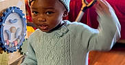 Tariyah joined the competition — help win amazing prizes! toddler, child, indoor, sweater, hat, pom_pom, toy, furniture, couch, carpet, wood_floor, person, face, smile, playful, curious, warm_lighting, clothing, hand, headwear