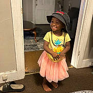 Tariyah joined the competition — help win amazing prizes! toddler, child, smiling, hat, bucket_hat, yellow_tshirt, pink_skirt, barefoot, indoor, doorway, carpet, slipper, table, furniture, happy, cute, person, portrait, home, clothing