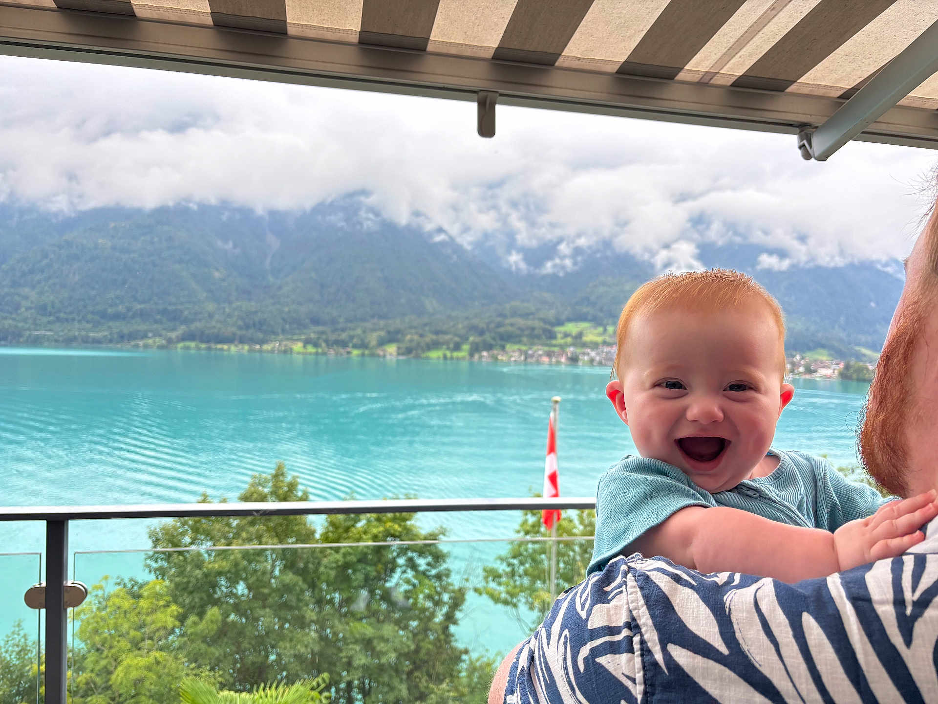 Stanley is registered to the contest to win money with this photo: baby, child, red_hair, smile, happy, adult, holding, outdoor, lake, mountains, water, trees, balcony, railing, awning, nature, scenic, person, portrait, summer