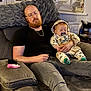 man, baby, sleeping, couch, living_room, glasses, beard, pacifier, hoodie, blanket, window, tissue_box, remote_control, phone, casual_clothing, home_interior, relaxation, seated, indoor, family