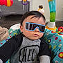 baby, infant, sunglasses, baby_seat, play_gym, toy, colorful, striped_clothing, hand, face, hair, indoor, floor, plush_toy, patterned_fabric, candid, portrait, cool, sitting, seat_cushion