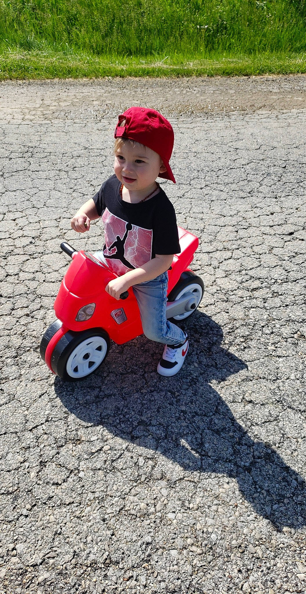 Arrow is registered to the contest to win money with this photo: asphalt, automotive_wheel_system, cap, carmine, child, driveway, fun, grass, headwear, helmet, person, recreation, riding_toy, road_surface, rolling, sitting, tire, toddler, toy, toy_vehicle