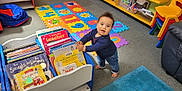 Elijah is registered to the contest to win money with this photo: baby, barefoot, books, bookshelf, carpet, child, childrens_books, colorful, education, floor, foam_mat, furniture, indoor, learning_area, playroom, portrait, smiling, standing, toddler, toy_chair