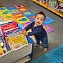 Elijah is registered to the contest to win money with this photo: toddler, child, bookshelf, books, foam_mat, colorful, carpet, barefoot, indoor, learning_area, toy_chair, childrens_books, standing, baby, playroom, education, floor, furniture, smiling, portrait