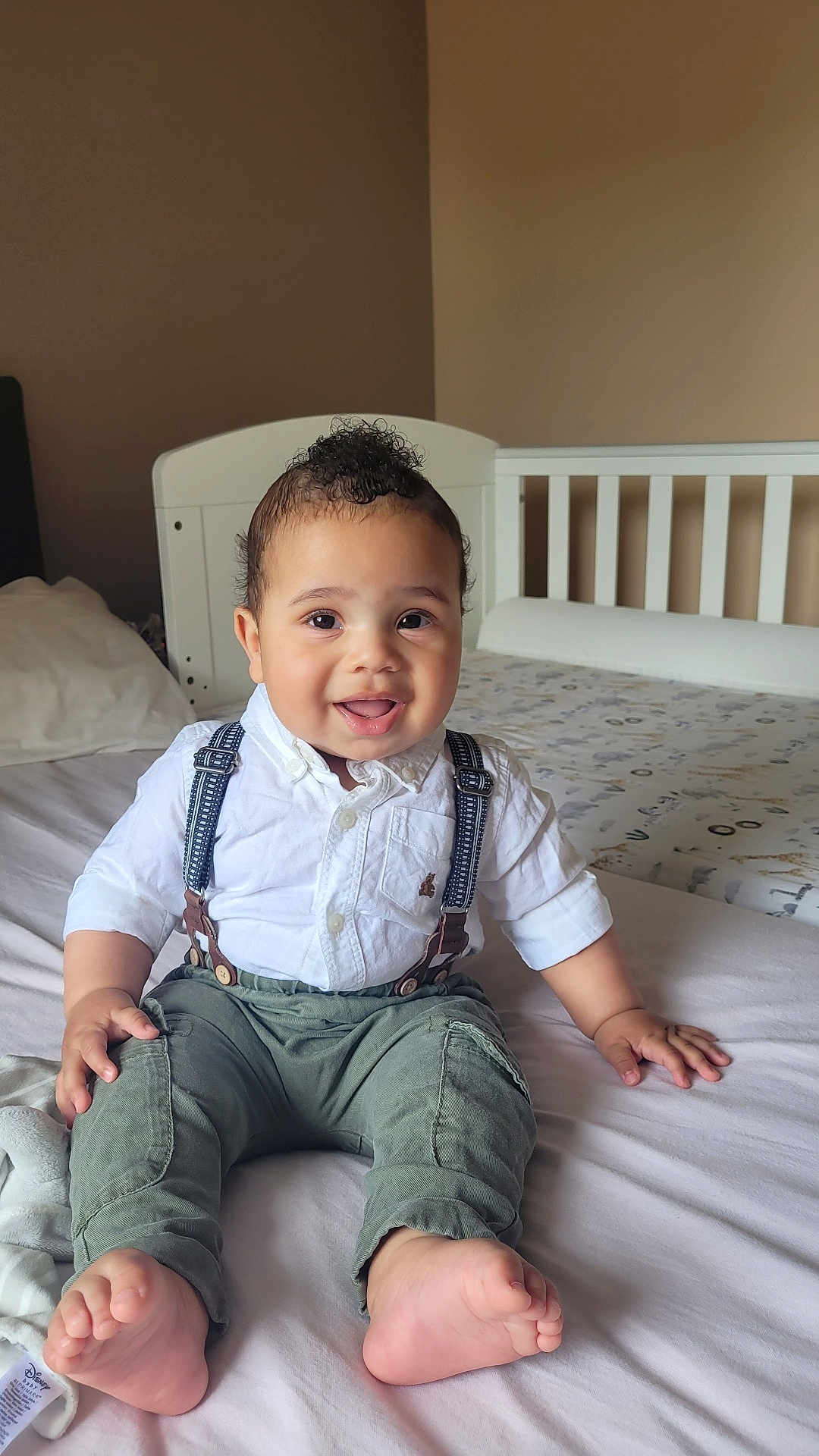 Elijah is registered to the contest to win money with this photo: baby, child, smiling, sitting, bed, shirt, suspenders, pants, feet, nursery, crib, curly_hair, happy, indoor, young_child, portrait, cute, infant, clothing, person