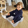 toddler, child, plastic_bin, socks, navy_blue_clothing, wooden_floor, indoor, furniture, chair, home, playful, sitting, person, floor, light, casual_clothing, cute, young_child, smiling, relaxed