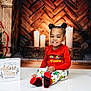 child, toddler, smiling, sitting, pajamas, red_shirt, patterned_pants, red_socks, hair_buns, fireplace, candles, holiday_decor, christmas_theme, wooden_background, decorative_sign, indoor, portrait, cute, cozy, smile