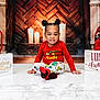 child, toddler, christmas, holiday, tree, decorations, gifts, presents, candles, fireplace, smile, sitting, red_shirt, patterned_pants, socks, poinsettia, ornaments, studio_backdrop, portrait, festive