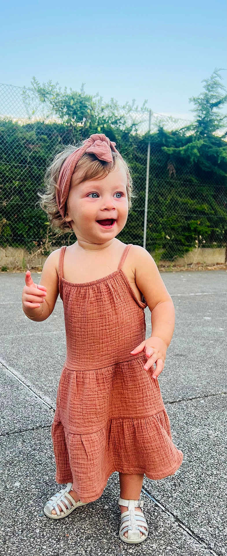 Cataleya participe au concours pour gagner de l'argent avec cette photo : toddler, child, smiling, curly_hair, headband, dress, outdoor, pavement, sandals, happy, person, young_child, summer, sunny, fence, greenery, playful, cute, portrait, daylight