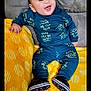 baby, child, smiling, blue_outfit, sitting, yellow_blanket, patterned_blanket, couch, happy, indoor, footwear, black_shoes, text_on_clothing, infant, portrait, cute, playful, cozy, young_child, face