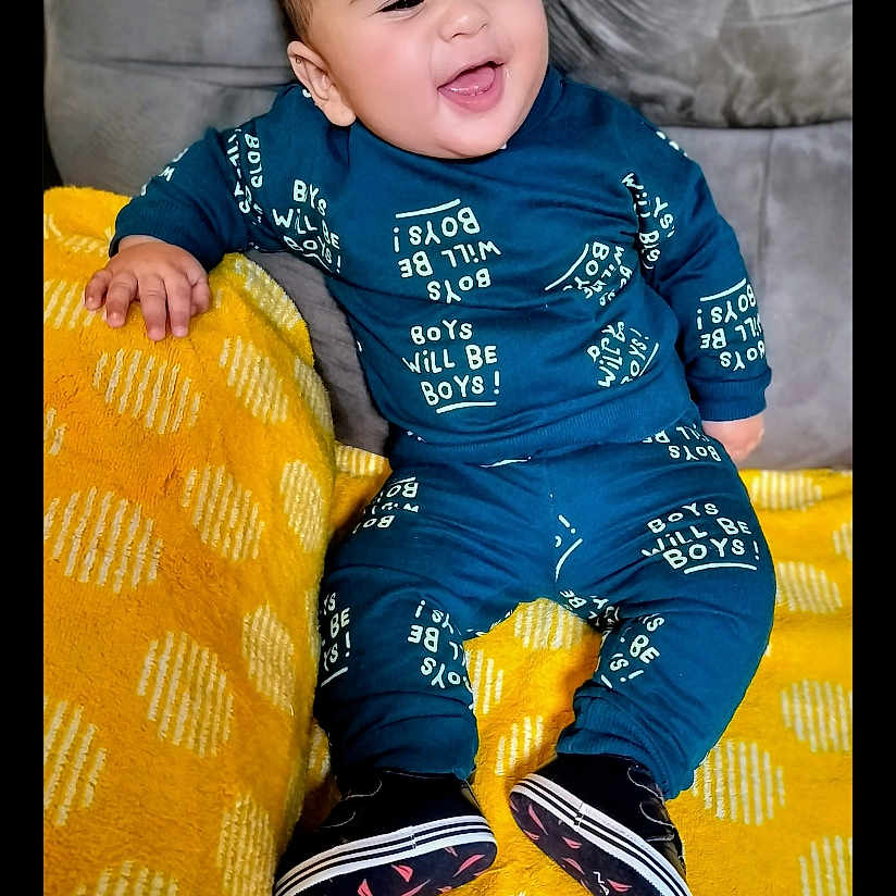 Chris Benítez joined the competition — help win amazing prizes! baby, black_shoes, blue_outfit, child, couch, cozy, cute, face, footwear, happy, indoor, infant, patterned_blanket, playful, portrait, sitting, smiling, text_on_clothing, yellow_blanket, young_child