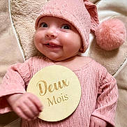 Maxine a rejoint le concours — aidez-le/la à gagner de superbes lots ! baby, infant, pink_clothing, knit_hat, pom_pom, blanket, smile, face, hand, wooden_plaque, milestone, two_months, lying_down, cute, soft_texture, portrait, young_child, happy, indoors, child