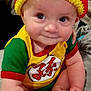 baby, infant, hat, knit_hat, pom_pom, colorful_clothing, yellow, red, green, smile, cute, sitting, bed, tattoo, arm, hand, foot, indoors, person, portrait