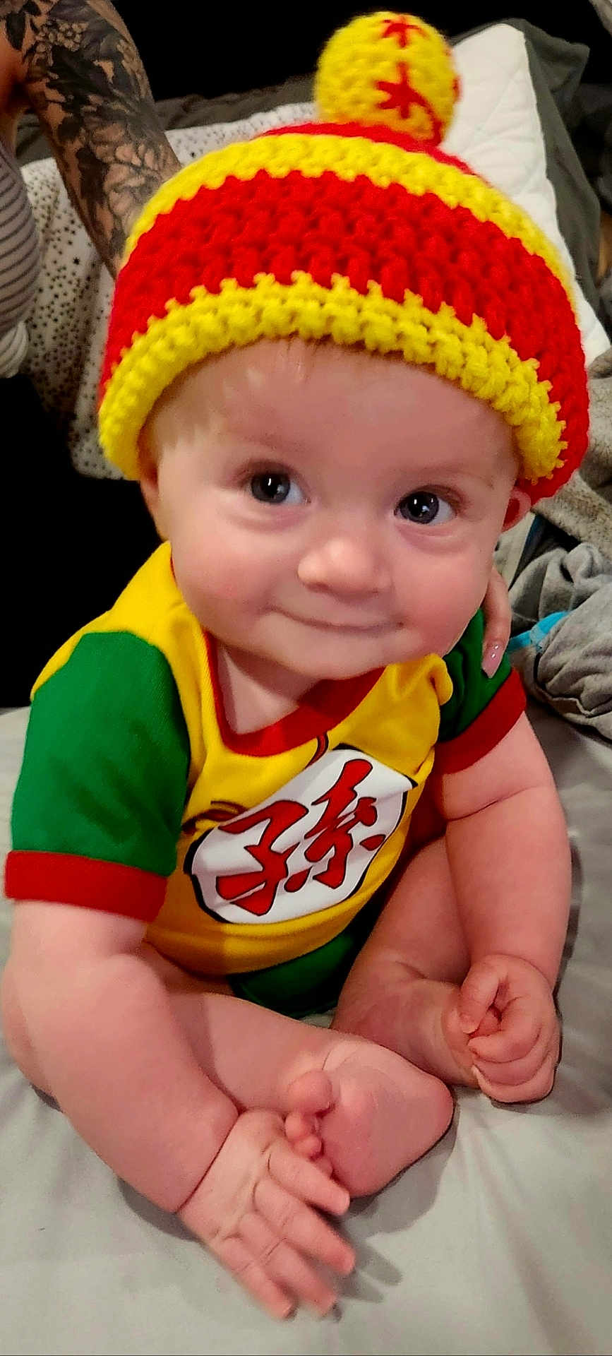 Victor joined the competition — help win amazing prizes! baby, infant, hat, knit_hat, pom_pom, colorful_clothing, yellow, red, green, smile, cute, sitting, bed, tattoo, arm, hand, foot, indoors, person, portrait