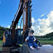 Kristyn is registered to the contest to win money with this photo: baby, child, excavator, construction_machine, outdoor, nature, grass, blue_sky, clouds, denim_overalls, barefoot, greenery, heavy_equipment, infant, portrait, vehicle, sunlight, field, cute, serious_expression