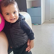 Télio participe au concours pour gagner de l'argent avec cette photo : toddler, child, smile, black_vest, indoor, person, sweater, toy, shelf, furniture, floor, hand, happy, cute, young_child, face, casual_clothing, room, light, portrait