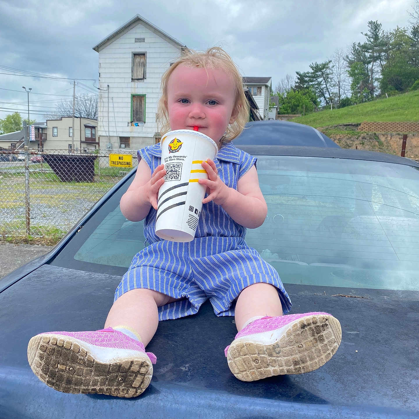Paris is registered to the contest to win money with this photo: toddler, child, car, drink, cup, blue_striped_outfit, pink_shoes, outdoor, cloudy_sky, grass, house, fence, red_straw, sitting, person, face, shoes, vehicle, daytime, young_child
