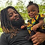 Charity is registered to the contest to win money with this photo: man, baby, child, smiling, holding, dreadlocks, beard, outdoor, nature, greenery, sunflower, black_clothing, portrait, happy, family, parent, infant, person, cute, affection