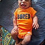Aubrey joined the competition — help win amazing prizes! baby, child, infant, smiling, laughing, orange_onesie, clothing, lying_down, quilt, patterned_blanket, happy, cute, skin, hand, foot, person, indoors, text_on_clothing, fabric, bright_color