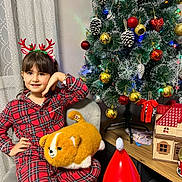 Shams is registered to the contest to win money with this photo: child, girl, smile, pajamas, plaid, reindeer_headband, plush_toy, stuffed_animal, christmas_tree, ornament, pinecone, bauble, christmas_decor, chair, curtain, indoor, holiday, festive, toy_house, santa_figure